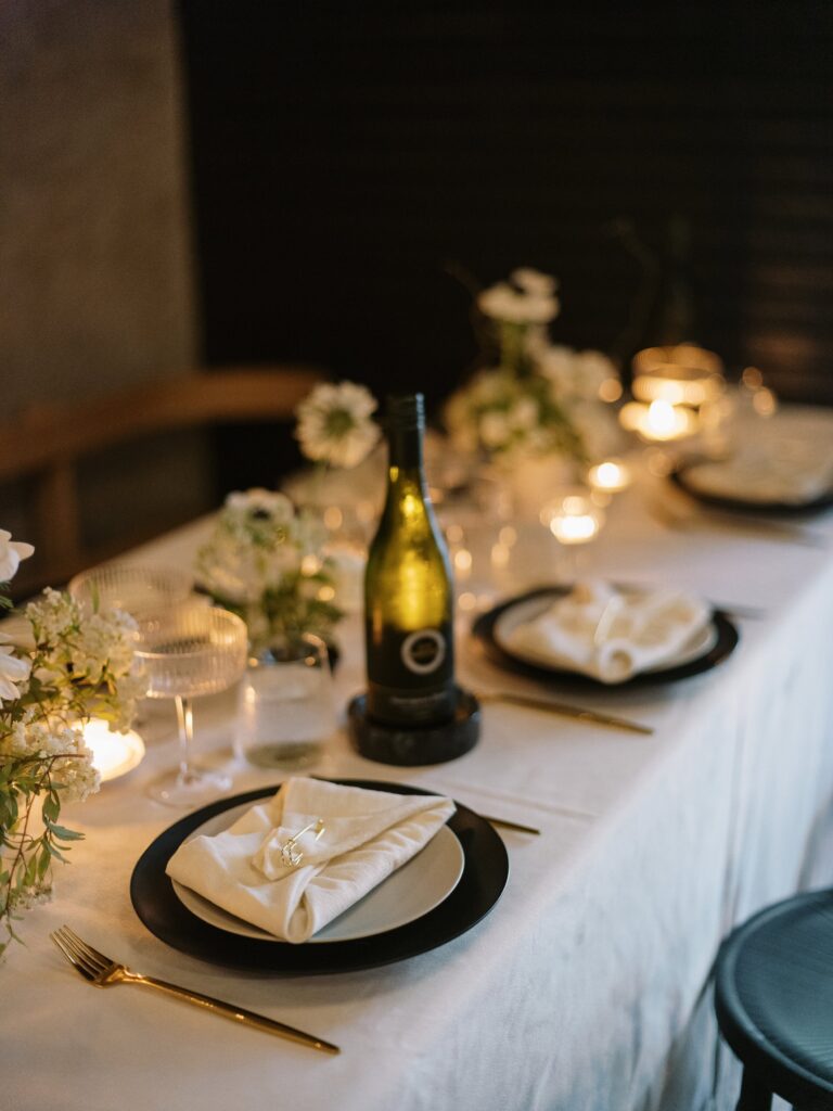A table set for a formal dinner with a white tablecloth, plates with folded napkins, a bottle of wine, candles, and floral decorations.