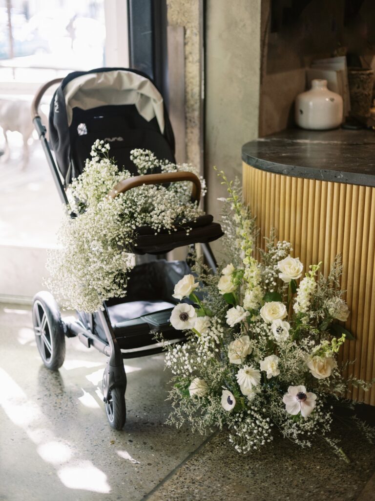 A stroller filled with delicate white flowers is positioned next to a floral arrangement on a countertop inside a room with sunlight streaming in.