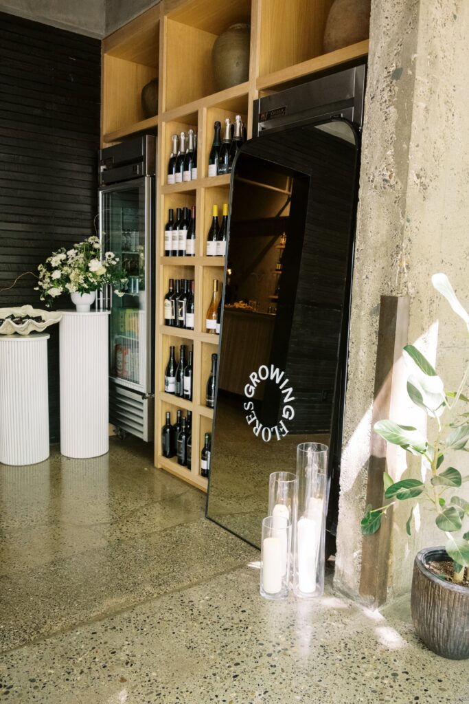 Wine bottles are displayed on wooden shelves next to a mirrored sign reading "GROWING OLD IS GETTING GORGEOUS," with decorative plants and tall candles nearby.