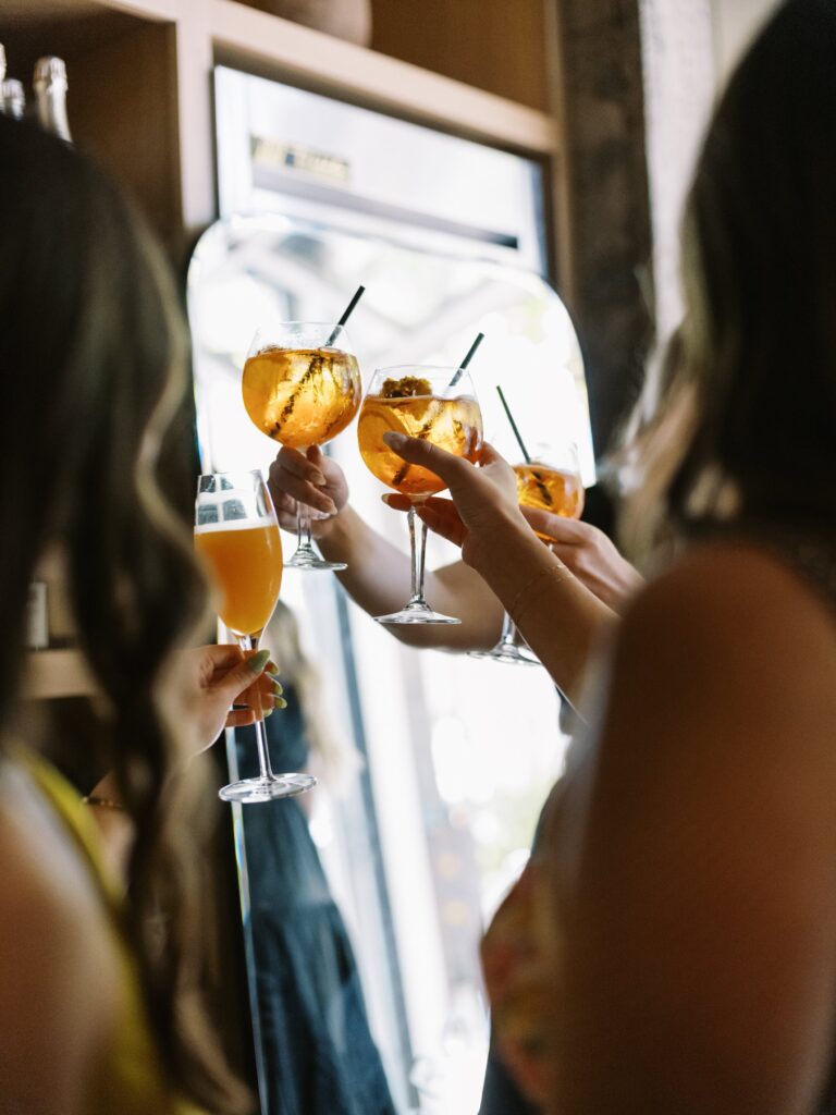 Four people clinking glasses filled with orange drinks in a celebratory toast near a mirror.