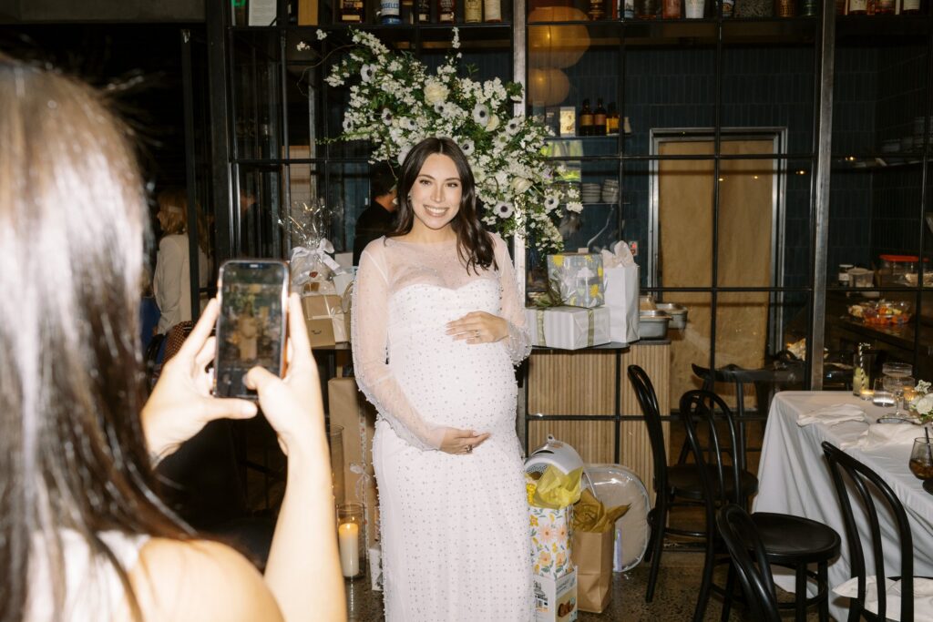A pregnant woman in a white dress poses for a photo at a celebration, surrounded by flowers and gifts. Another person takes her picture with a phone.