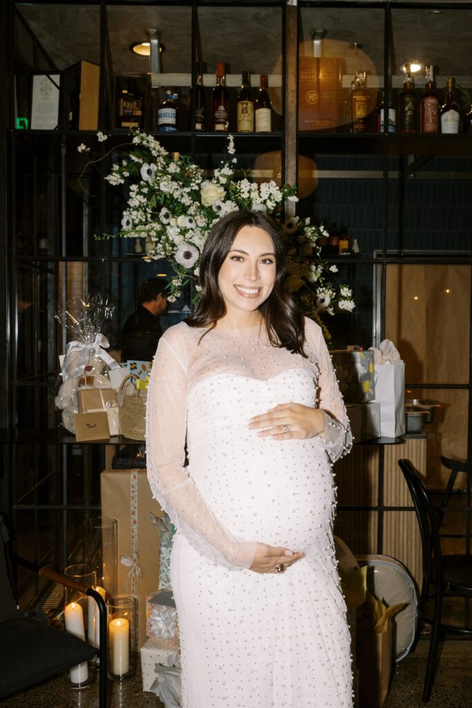 A pregnant woman in a white dress stands smiling in front of a decorated area with flowers and gifts.
