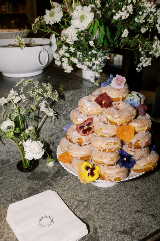 A stack of sugar-coated donuts adorned with edible flowers is displayed on a plate beside a small bouquet and napkins on a concrete countertop.