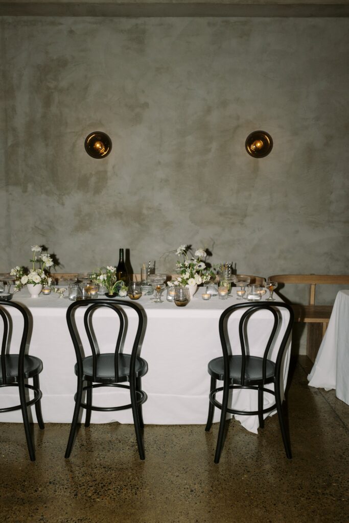 A simple, elegant dining table setup with white tablecloth, black chairs, floral centerpieces, and tableware against a muted gray wall with two round wall lights.