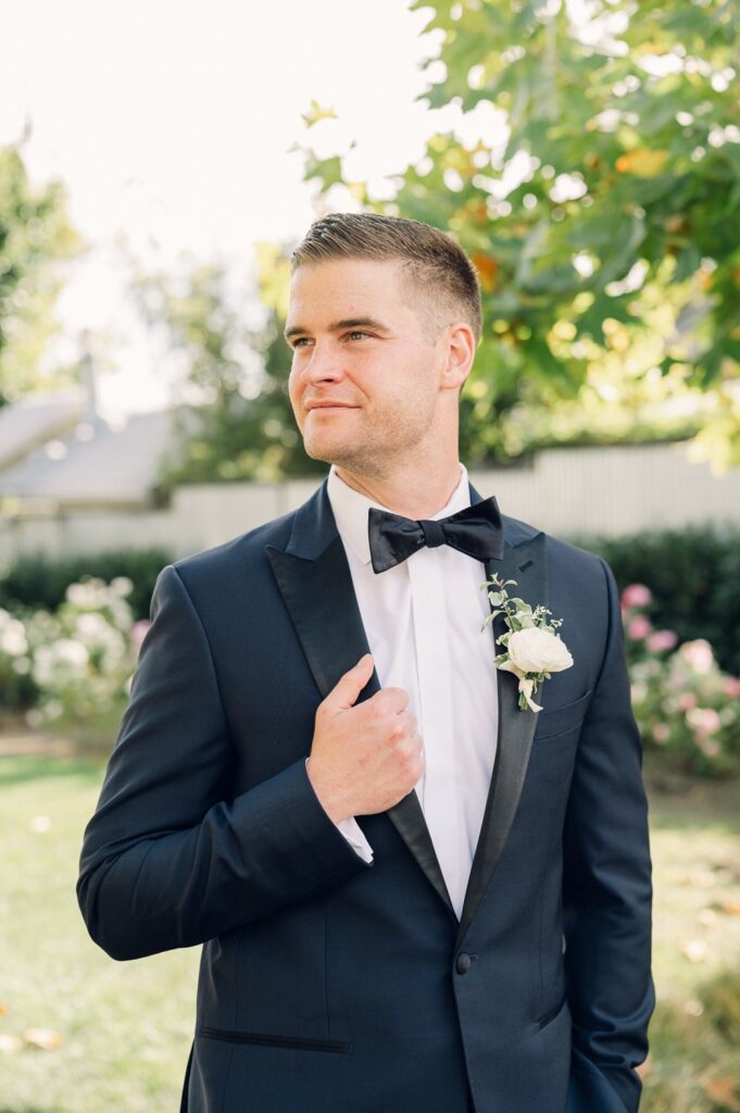 A man in a tuxedo, holding his lapel, stands outdoors with greenery and flowers in the background.