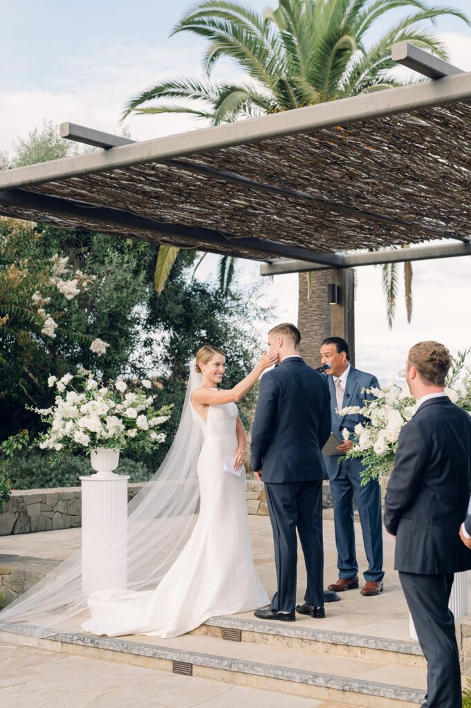 A bride and groom stand at an outdoor altar during their wedding ceremony, with a wedding officiant and a groomsman nearby. Lush greenery and white flowers surround the setting.