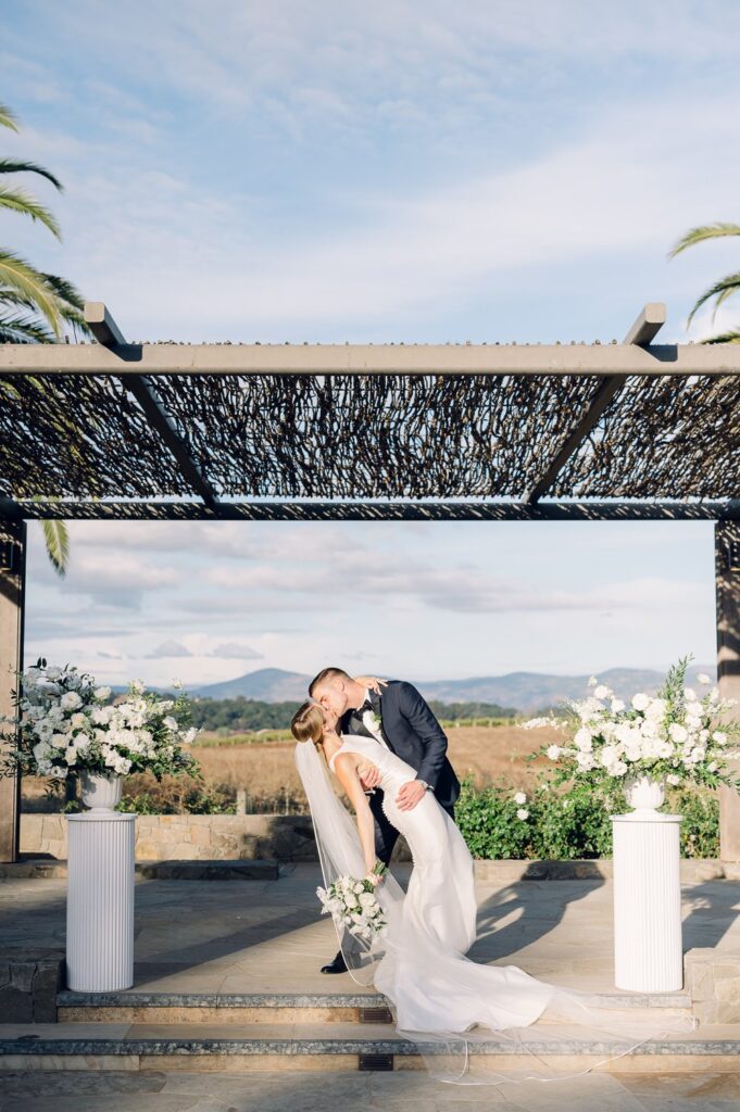 A couple shares a kiss under a pergola during their wedding ceremony, surrounded by floral arrangements and overlooking a scenic landscape.