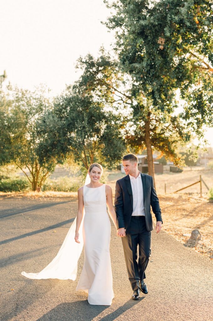 A couple in formal attire walks hand in hand on a sunlit path, surrounded by trees.