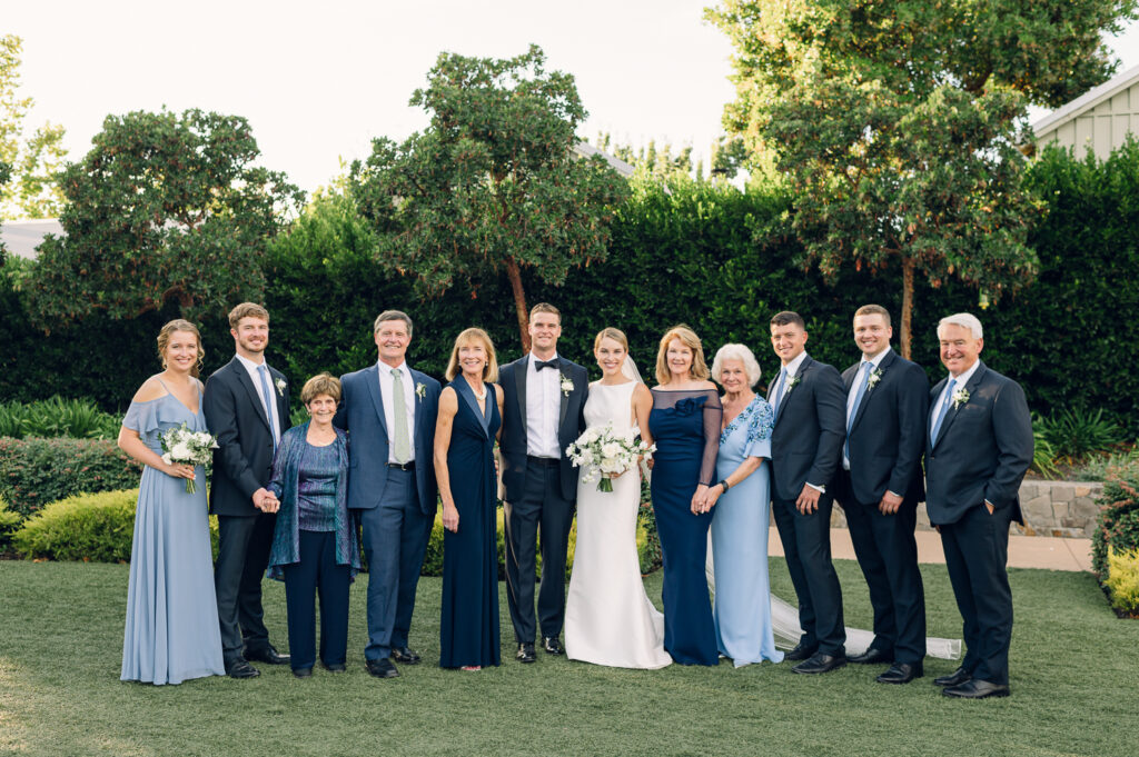 A group of twelve people, dressed formally, stand outdoors on the lush Napa grass. Two in the center are holding flower bouquets. Majestic trees and a neatly trimmed hedge serve as the backdrop for this lovely wedding scene.