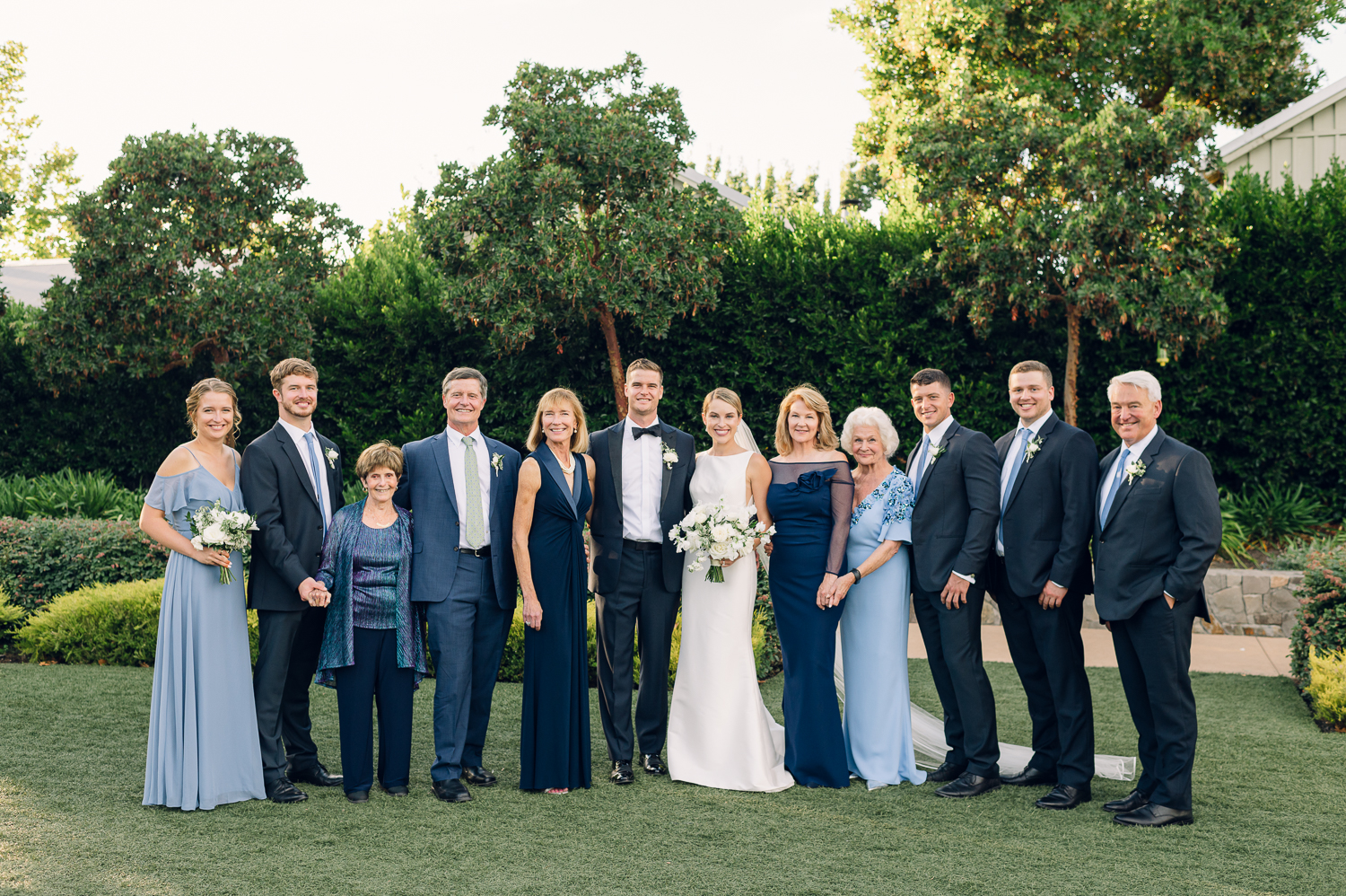 A group of twelve people, dressed formally, stand outdoors on the lush Napa grass. Two in the center are holding flower bouquets. Majestic trees and a neatly trimmed hedge serve as the backdrop for this lovely wedding scene.