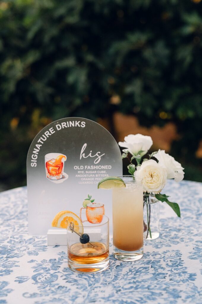 Two cocktails on a floral tablecloth next to a glass vase with white flowers and a sign reading "Signature Drinks: His Old Fashioned.
