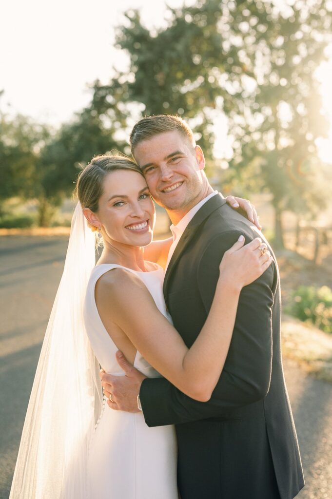 A bride and groom embrace and smile at the camera on a sunlit outdoor path, with trees in the background.