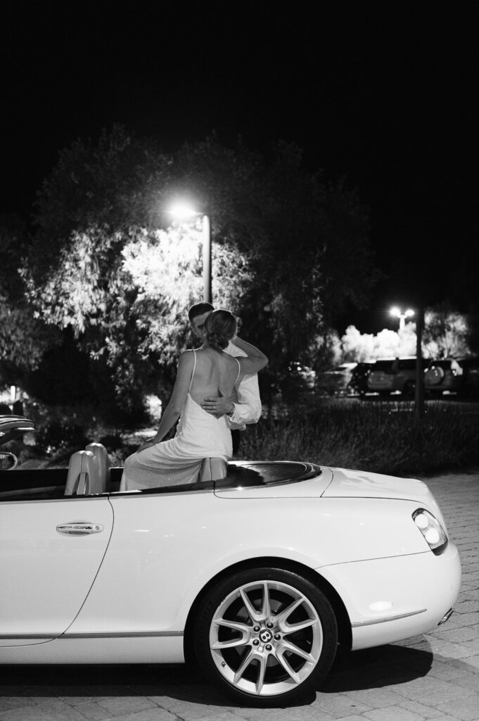 A couple embraces while sitting on the back of a white convertible at night, under streetlights.
