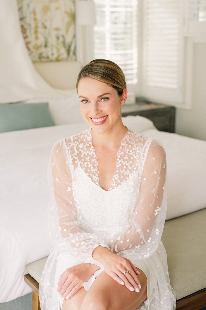 A woman in a white, sheer, embroidered dress sits on a bench in a bright room, smiling at the camera.