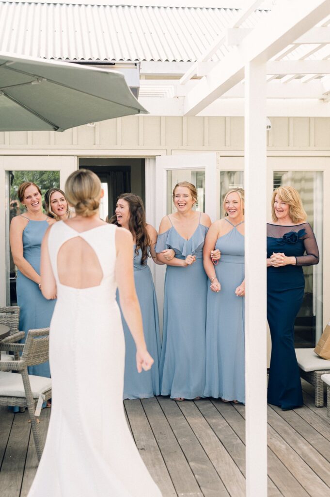 A bride walks toward a group of smiling bridesmaids in blue dresses on a wooden deck.