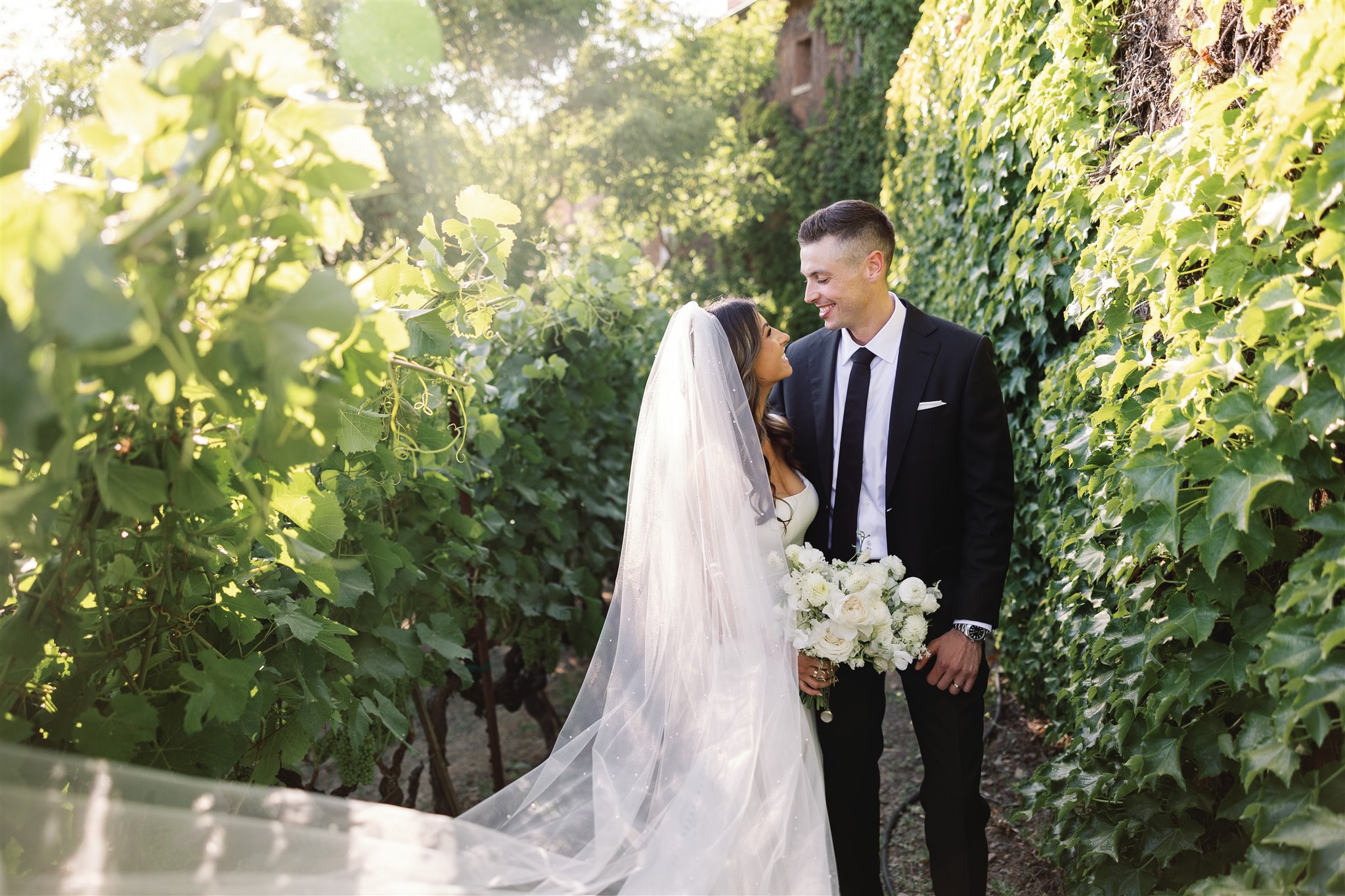 In a sunlit garden reminiscent of an Italian-inspired wedding, the bride and groom stand close together, surrounded by lush green foliage. The bride holds a bouquet of white flowers, adding to the romantic ambiance.