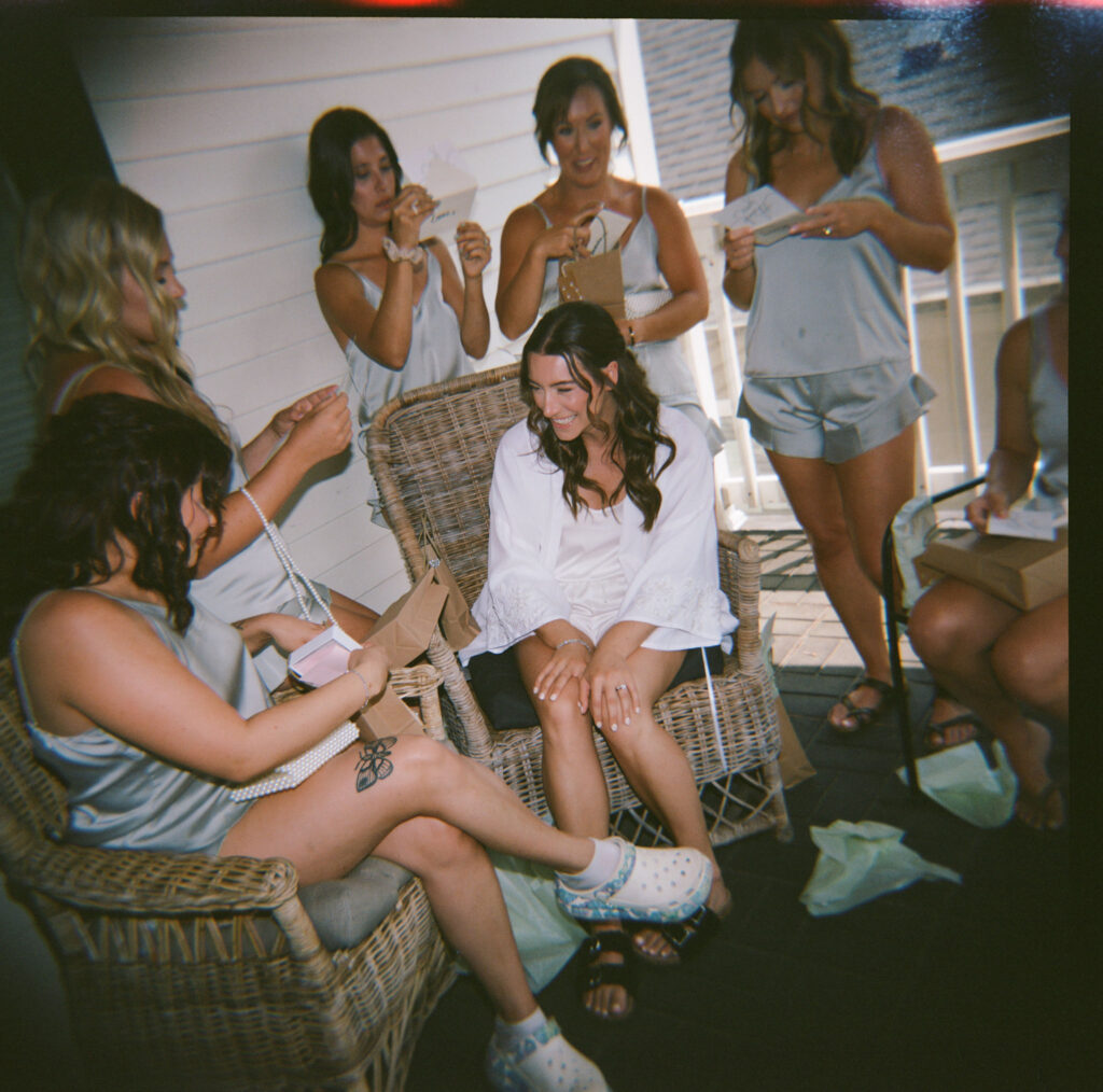 A group of women in casual attire sit and stand on a porch, sharing a light moment and exchanging cards or notes.