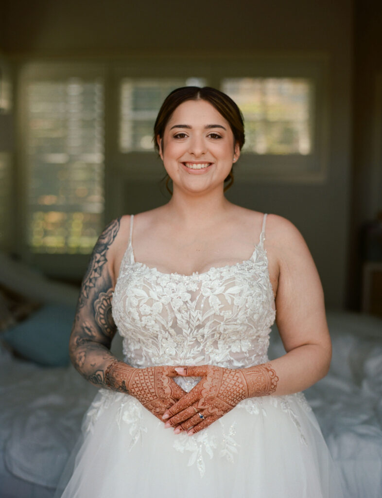 A bride in a white lace wedding dress with henna on her hands and a tattoo on her arm stands indoors, smiling.