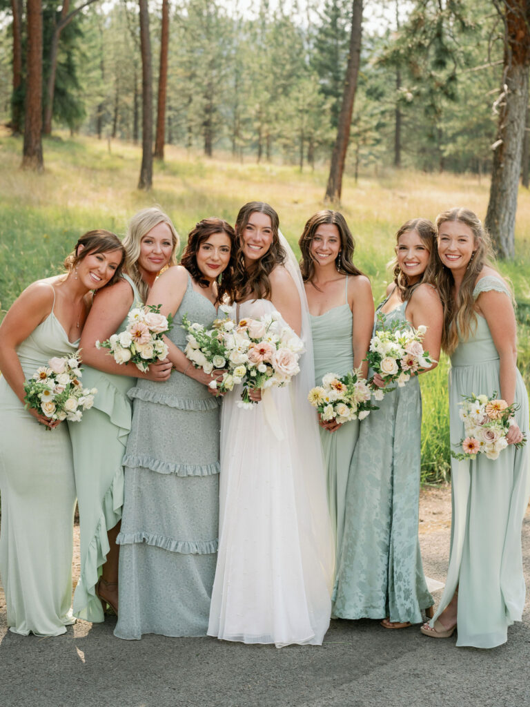 A bride and six bridesmaids in light green dresses smile and hold bouquets in a forest setting.