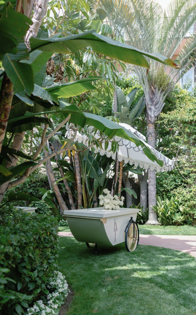 A white cart with flowers underneath a striped umbrella, surrounded by lush green tropical plants.