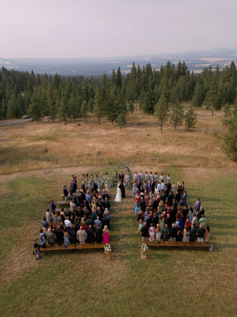 Aerial view of an outdoor wedding ceremony in a field surrounded by trees. Guests are seated on each side of the aisle as the couple stands at the altar.