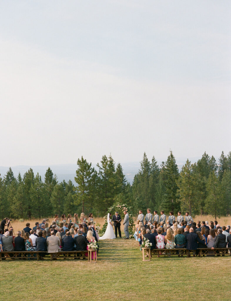 Outdoor wedding ceremony with guests seated on wooden benches, facing a couple standing at an altar in a forested area. Bridesmaids and groomsmen are lined up on either side.