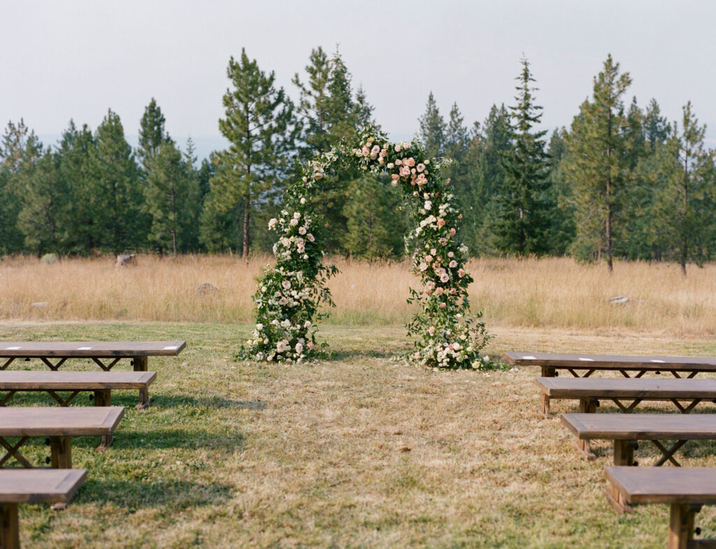 A floral wedding arch set in an outdoor field with wooden benches on either side, surrounded by trees.