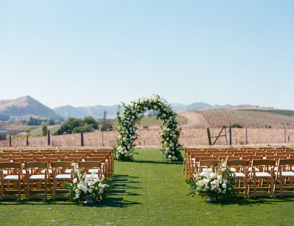 Outdoor wedding setup with wooden chairs facing a floral archway on a grassy field, surrounded by scenic hills and clear sky.