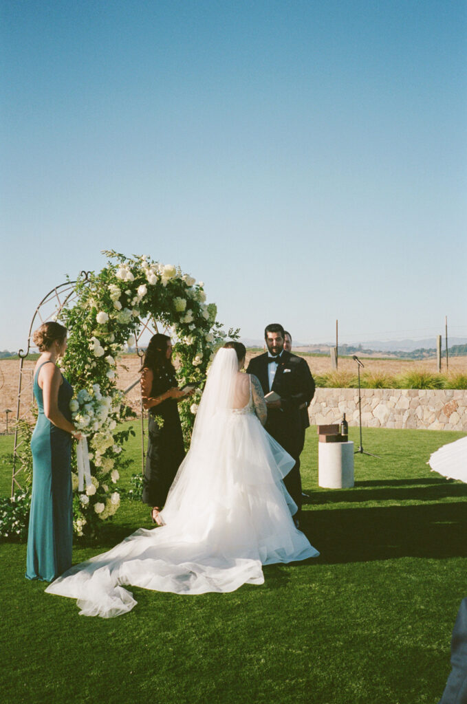 A bride and groom stand at an outdoor wedding ceremony. A person officiates under a floral arch while a bridesmaid in a blue dress stands nearby.