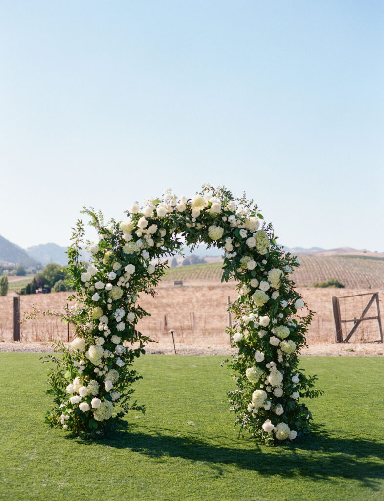A floral arch made of white flowers and greenery stands on a grassy field with a rural landscape and clear blue sky in the background.