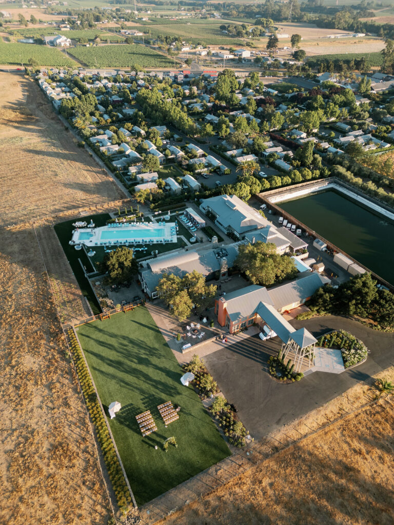 Aerial view of a rural landscape with a cluster of buildings, a rectangular pool, a green field, and surrounding farmland stretching into the distance.