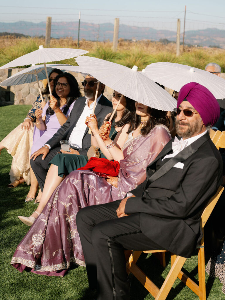 People seated outdoors, dressed formally, holding white parasols under sunny skies.