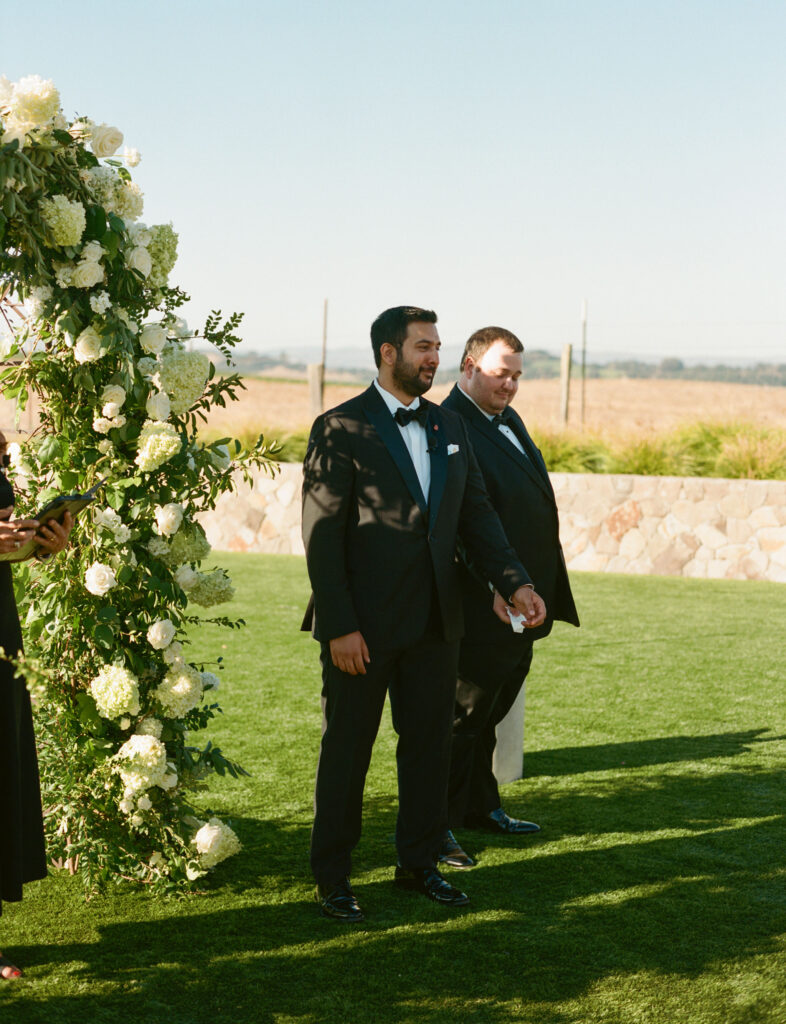Two men in formal suits stand on a grassy field beside a floral arch, holding hands under a clear sky.