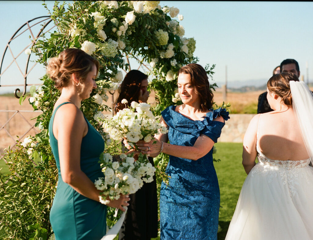 A wedding ceremony with women in elegant dresses near a floral arch. One woman holds a bouquet, another adjusts flowers. A bride stands with her back to the camera.