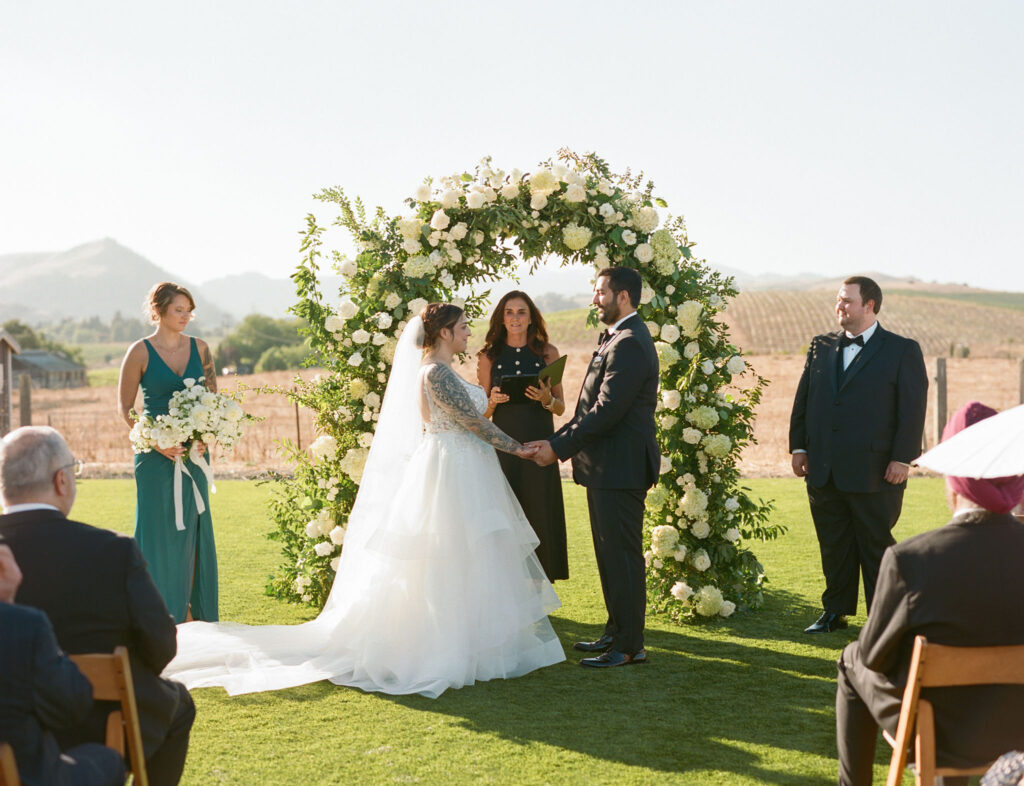 A couple stands before an officiant under a flower arch during an outdoor wedding ceremony, with two people standing beside them.