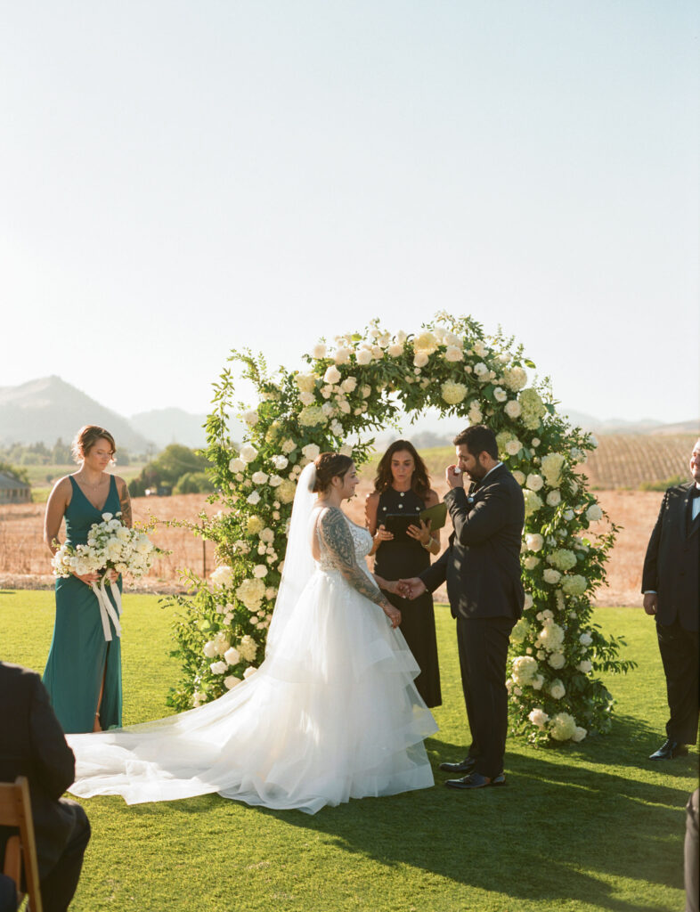 A wedding ceremony outdoors with a bride and groom standing under a floral arch. The bride is in a white dress, and the groom is in a dark suit. A bridesmaid and an officiant are present.