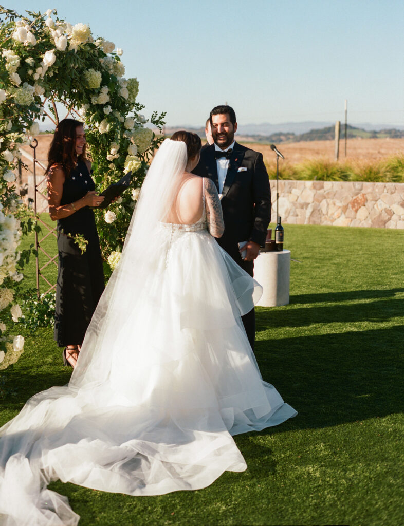 A couple stands before an officiant during an outdoor wedding ceremony. The bride wears a white gown with a long veil, and the groom is in a tuxedo. A floral arch is in the background.