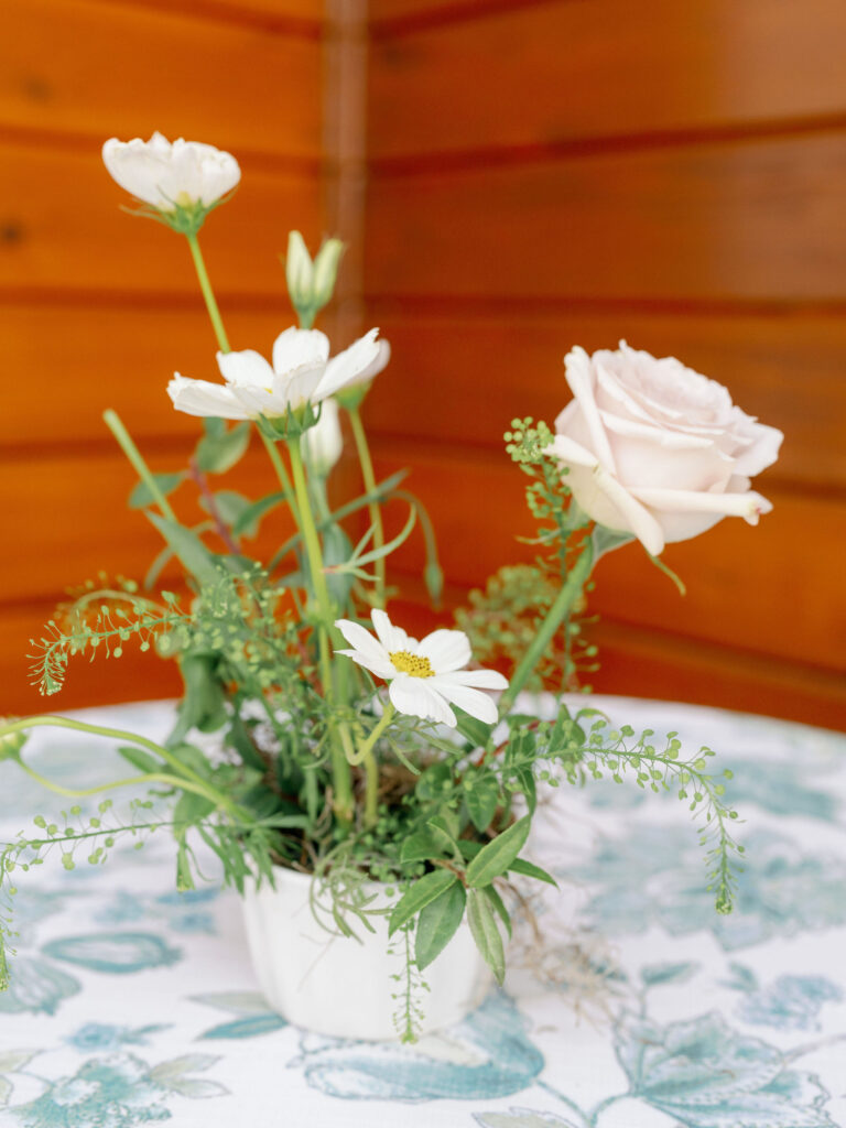 A small floral arrangement with white roses and daisies in a white vase, placed on a patterned tablecloth against a wooden wall background.