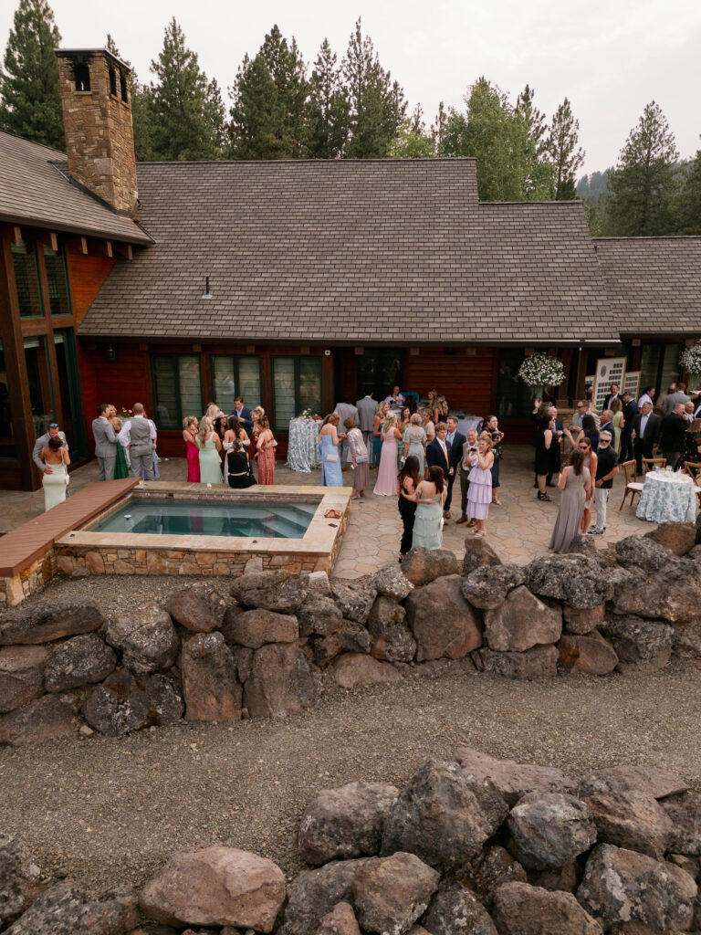 A group of people gather on a patio near a small pool outside a large wooden building surrounded by trees.