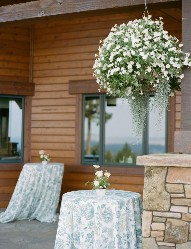 Hanging floral arrangement and tables covered with floral-patterned cloth outside a wooden building with stone column.