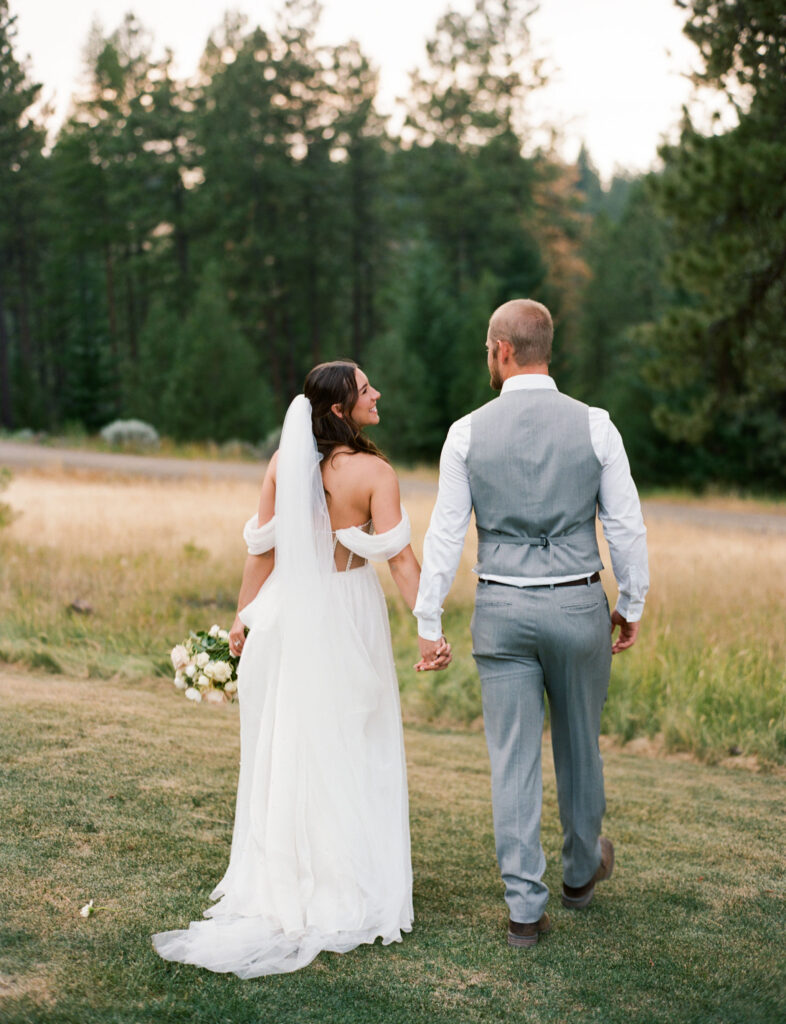 A bride and groom hold hands and walk together on a grassy field with trees in the background.