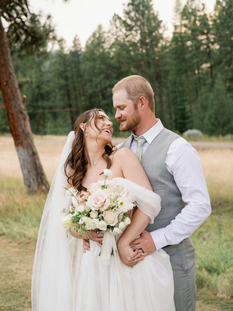 A bride and groom smile at each other while standing in a field, surrounded by trees. The bride holds a bouquet of white and pink flowers.