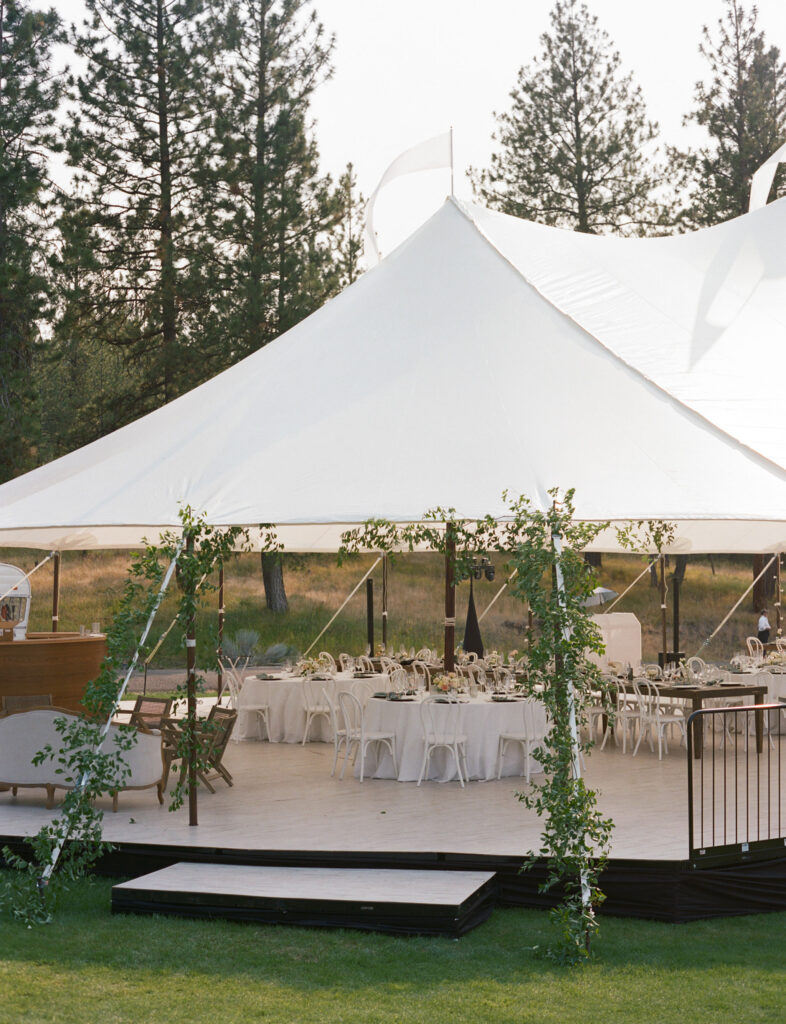 White tent set up outdoors with round tables and chairs, decorated with greenery, on a wooden platform with steps. Trees visible in the background.