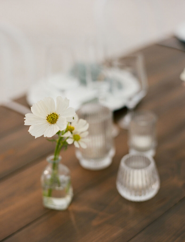 A small bouquet of white flowers in a glass vase sits on a wooden table surrounded by several empty glass cups.