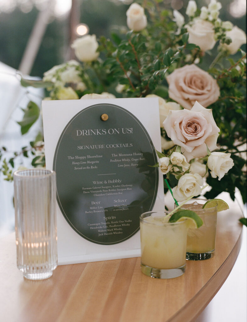 Menu with drink options and cocktails next to a floral arrangement and two glasses with drinks on a table.