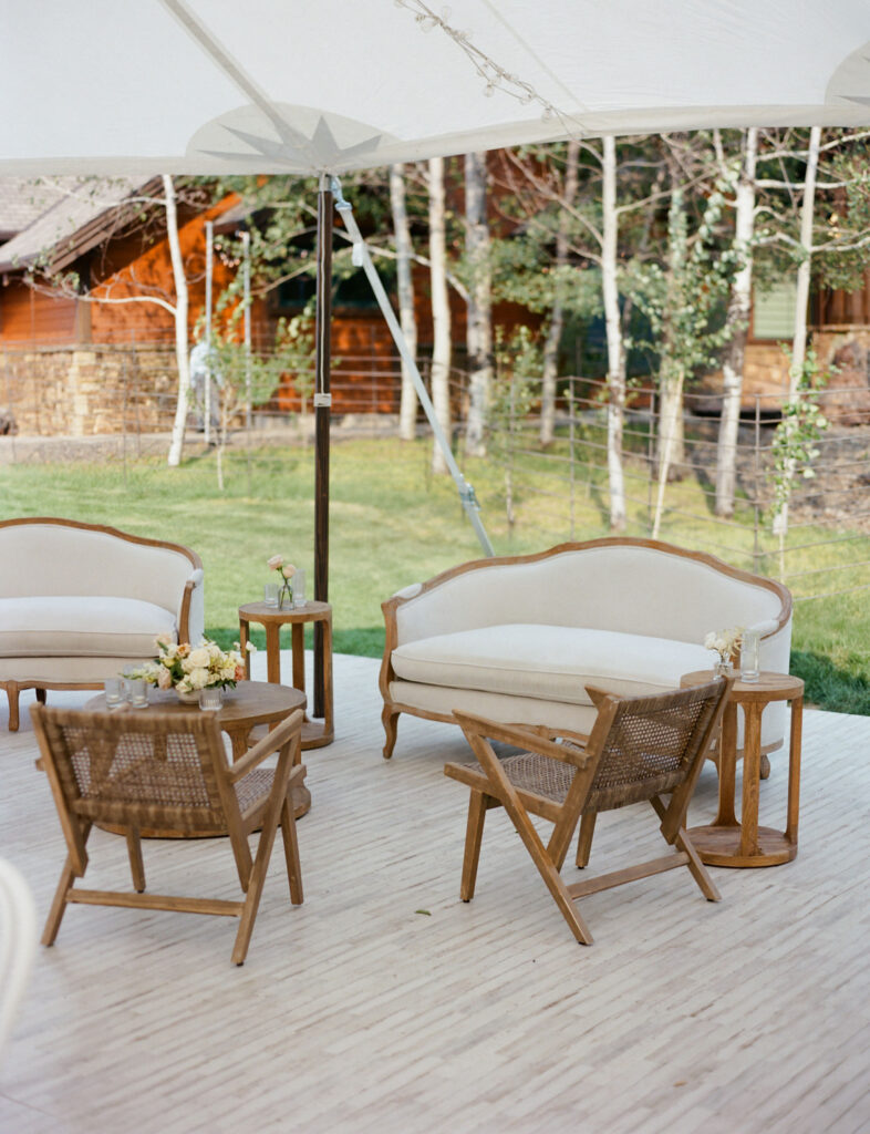 Outdoor seating area with wooden and upholstered chairs, side tables, and floral arrangements under a canopy. Background includes trees and a rustic building.