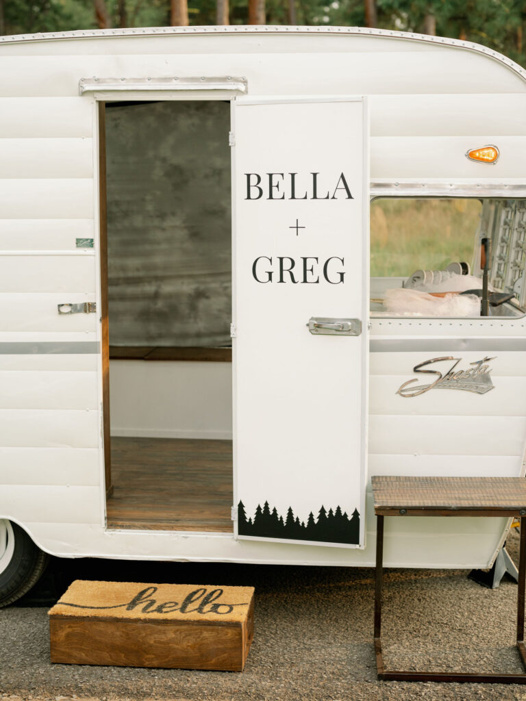 White vintage trailer with an open door displaying "Bella + Greg." A "hello" mat lies at the entrance with a step beneath it.