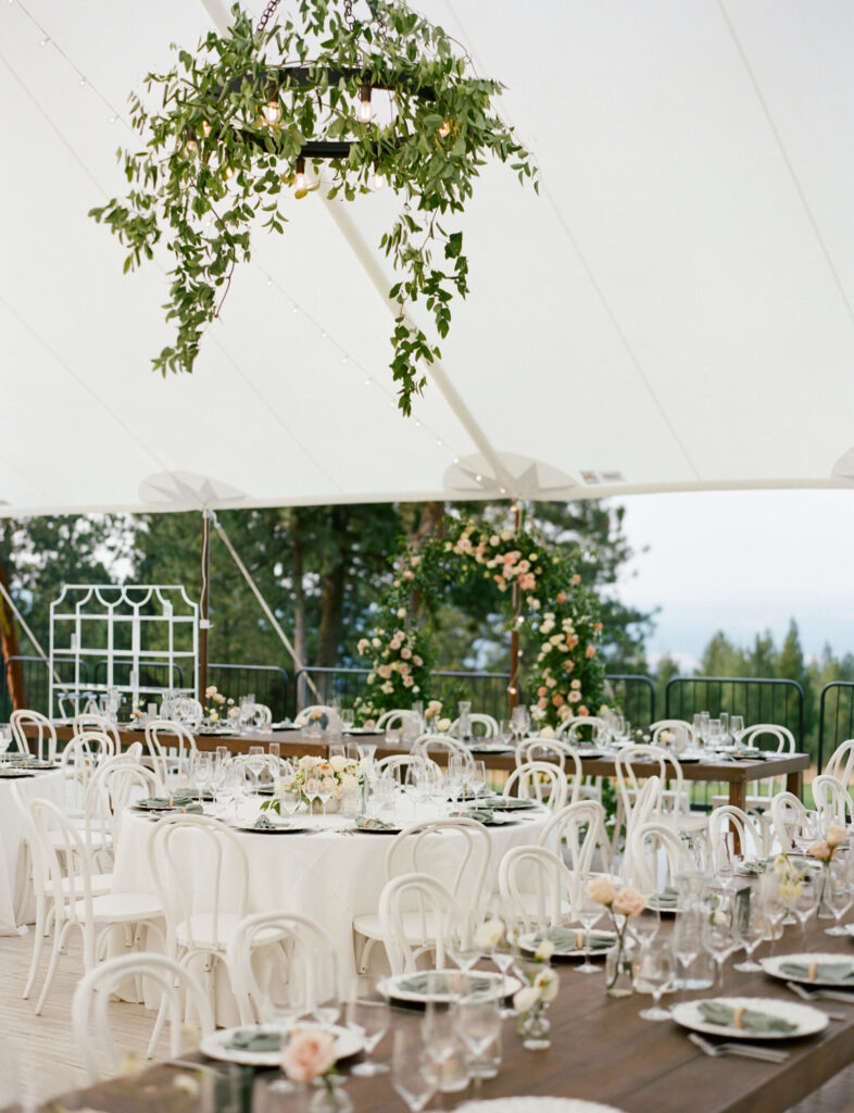 Elegant outdoor wedding reception under a tent, featuring round and rectangular tables with white chairs, floral centerpieces, and a hanging greenery arrangement.
