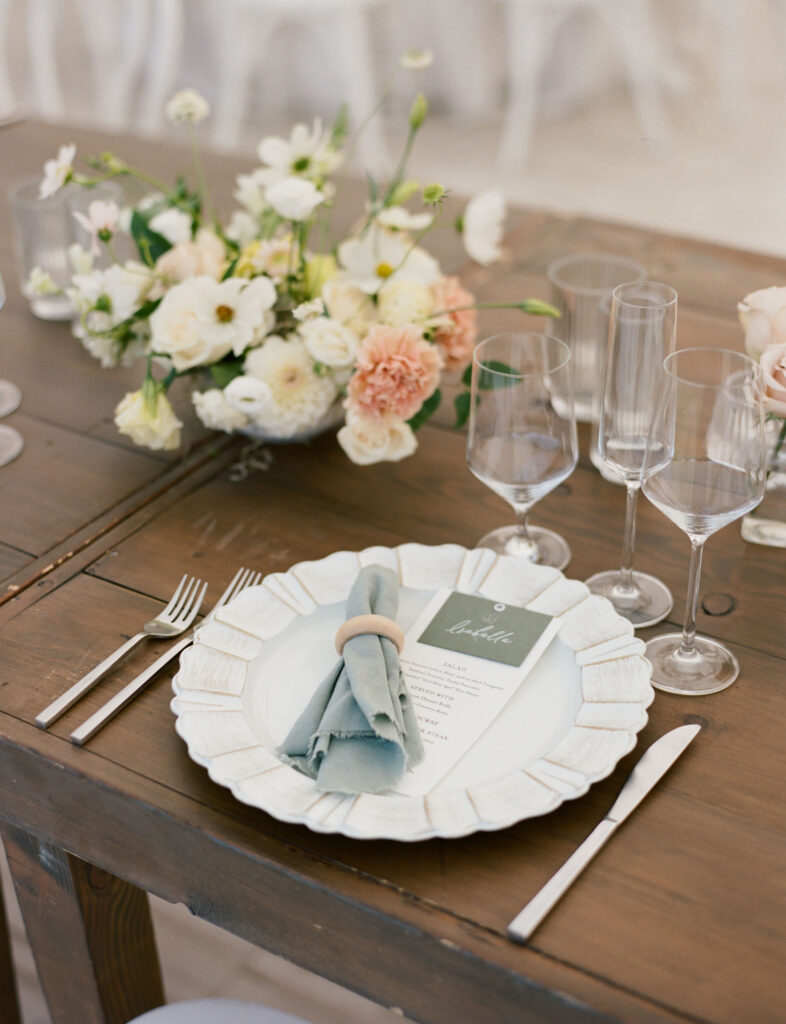 Elegant table setting with a floral centerpiece, neatly folded napkin on a plate, menu card, and empty wine glasses on a wooden table.