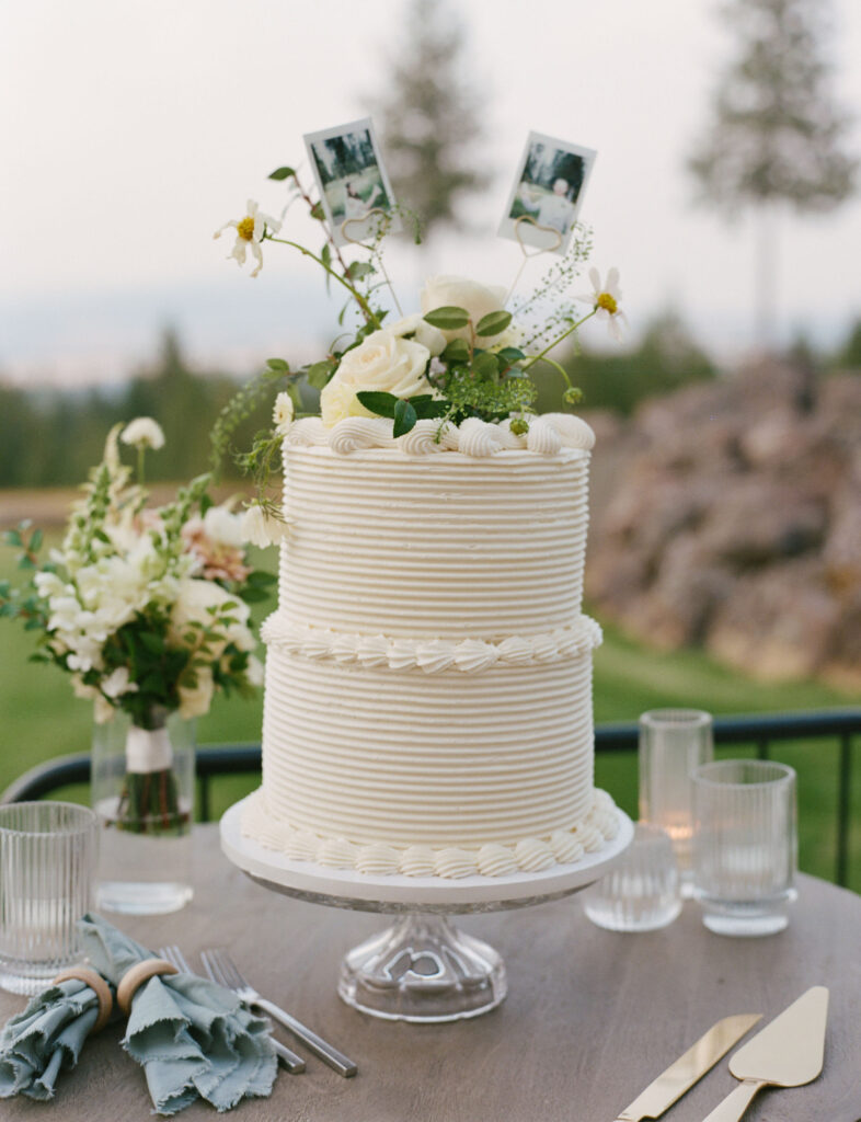 A two-tier white cake with floral decorations and photo toppers is on a table with glassware, cutlery, and floral arrangements, set outdoors with trees and rocks in the background.